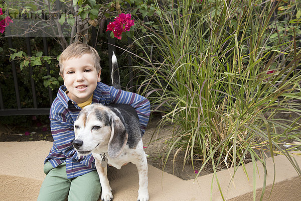 Porträt eines Jungen  der auf einer Parkwand sitzt und den Hund umarmt.