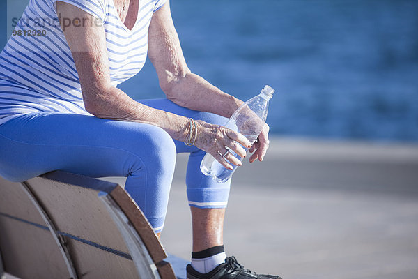 Seniorin sitzt auf einer Bank am Meer und hält Wasser in Flaschen.