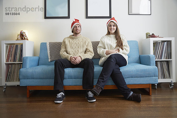 Porträt eines jungen Paares mit Weihnachtsmützen auf dem Sofa im Wohnzimmer