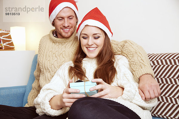 Junges Paar mit Weihnachtsmützen auf dem Sofa mit Blick auf das Weihnachtsgeschenk