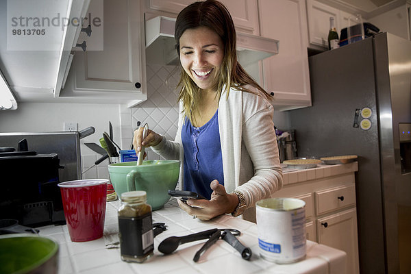 Frau bereitet Essen in der Küche zu  während sie das Rezept vom Smartphone liest.