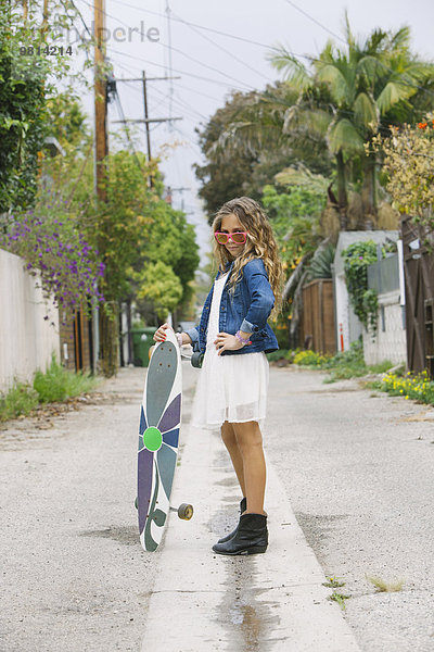 Mädchen steht auf der Straße und hält ein Skateboard.