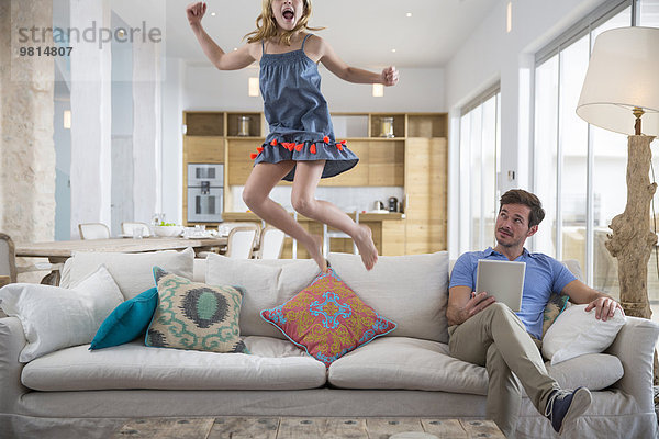 Mädchen springt in der Luft vom Wohnzimmersofa  während Vater ein digitales Tablett benutzt.