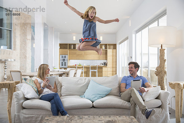 Mädchen springen in der Luft vom Wohnzimmersofa  während die Eltern ein digitales Tablett benutzen.