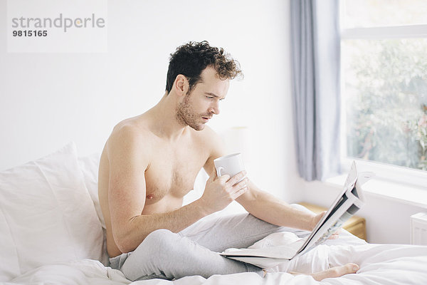Junger Mann im Bett liegend  Kaffee trinkend und Zeitung lesend
