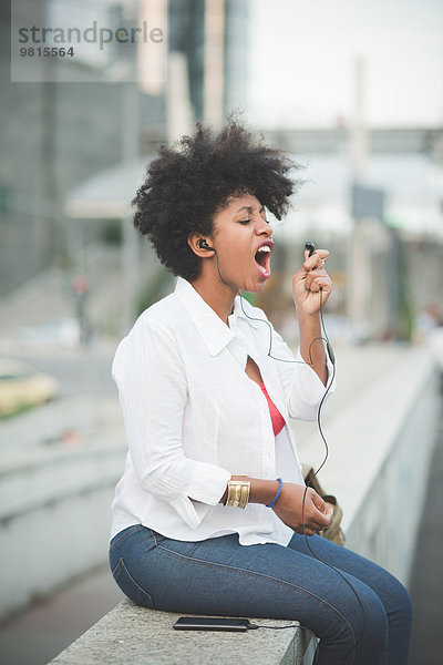 Junge Frau singt in den Kopfhörer  während sie auf der Wand des Einkaufszentrums sitzt.