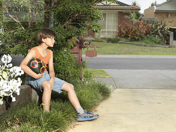Mürrischer Junge sitzt auf einer Vorstadtmauer und hält einen Fußball.