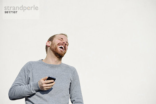Porträt eines lachenden jungen Mannes mit Smartphone vor weißem Hintergrund