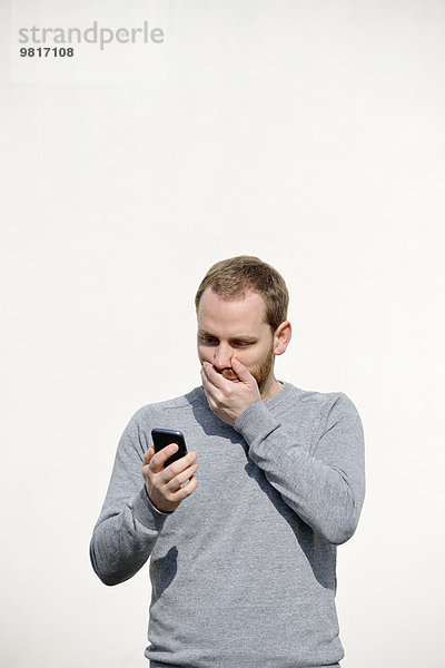 Porträt eines schockierten jungen Mannes  der sein Smartphone vor weißem Hintergrund ansieht