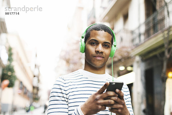 Spanien  Barcelona  Portrait eines lächelnden jungen Mannes  der Musik mit grünen Kopfhörern auf der Straße hört.