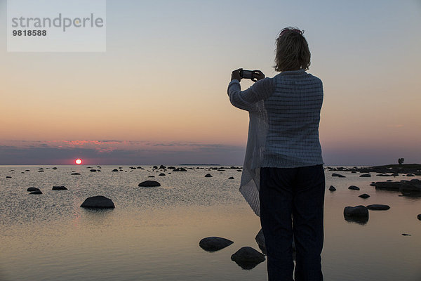 Estland  Kaesmu  Frau beim Fotografieren des Sonnenuntergangs mit Smartphone