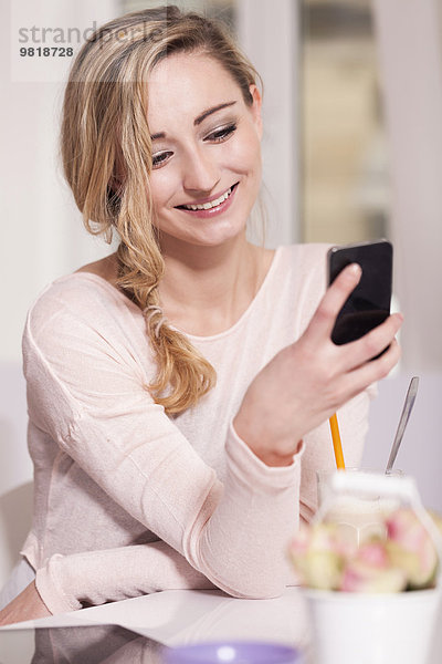 Porträt einer lächelnden blonden Frau  die mit dem Smartphone in einem Café sitzt.