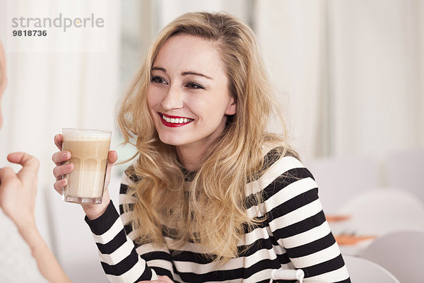 Lächelnde blonde Frau sitzt in einem Kaffee und hält ein Glas Latte Macchiato.