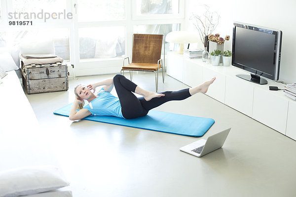Frau mit Laptop auf Turnmatte im Wohnzimmer