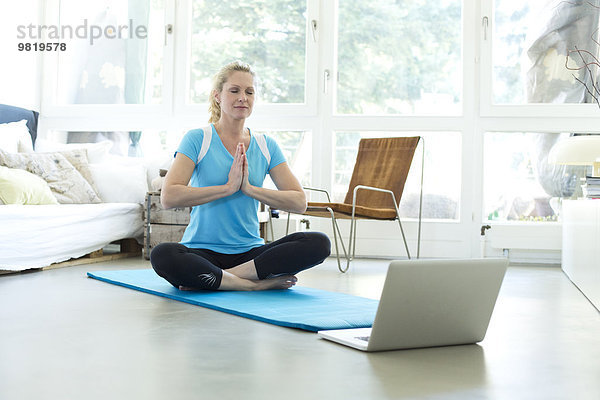 Frau mit Laptop praktiziert Yoga auf Turnmatte im Wohnzimmer