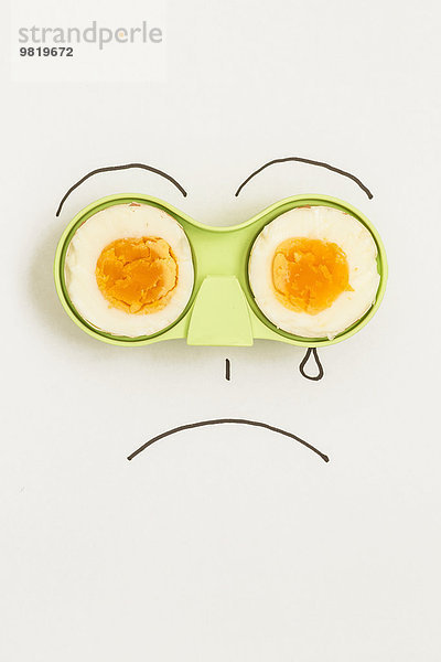Zwei Hälften eines Eies in grünem Halter mit traurigem Gesicht drumherum.