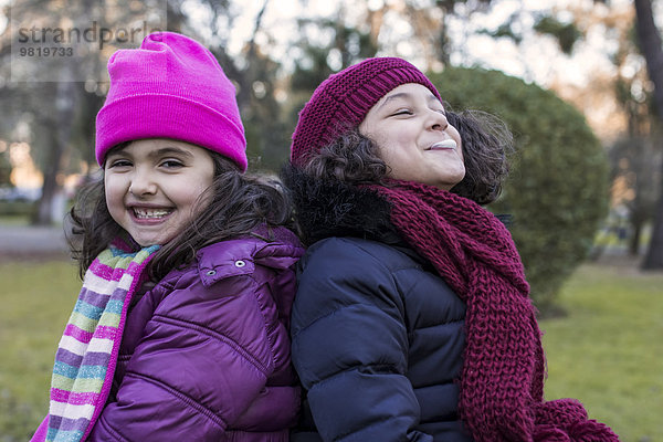 Portrait von zwei glücklichen kleinen Mädchen Rücken an Rücken in einem Park an einem Wintertag