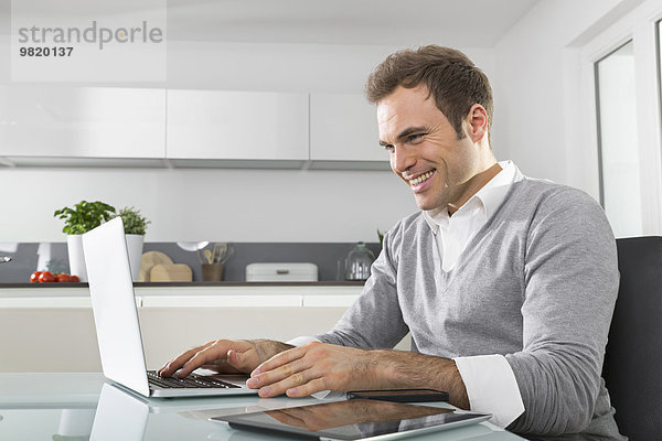 Lächelnder Mann sitzt in der Küche mit Laptop