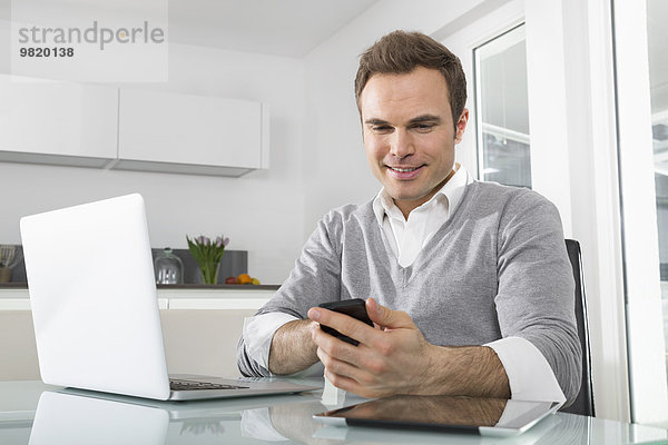 Lächelnder Mann sitzend in der Küche mit Laptop über Smartphone