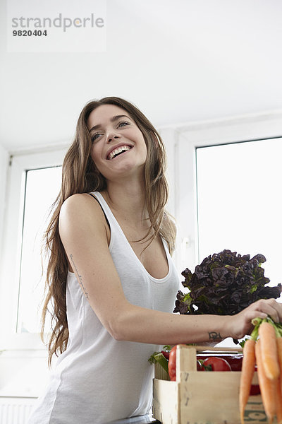 Fröhliche junge Frau in der Küche mit Kiste voller frischem Gemüse