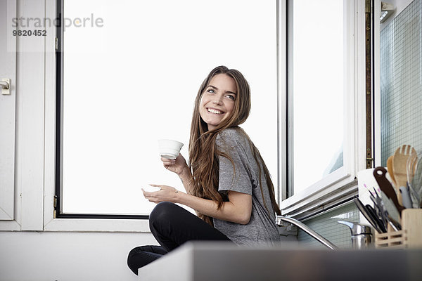 Junge Frau in der Küche sitzt am Fenster und trinkt Kaffee.