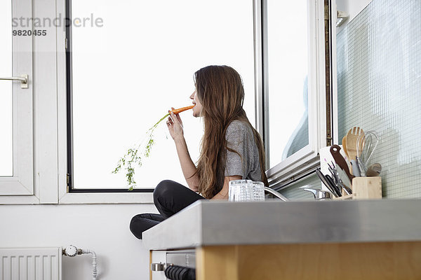 Junge Frau in der Küche sitzt am Fenster und isst Karotten.