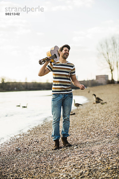 Mann steht am Flussufer und hält die Gitarre.