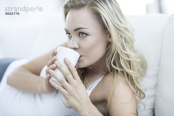 Blonde Frau liegt auf einer Couch und trinkt Kaffee.