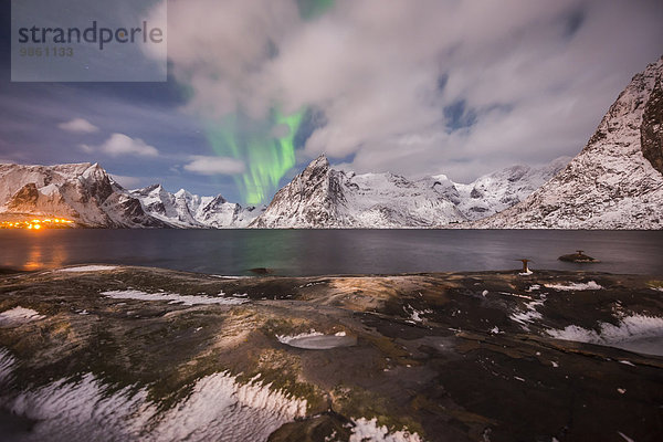 Aurora borealis mit Wolken  Felsen am Meer  eine kleine Stadt und Berge hinten  Insel Hamnoy  Lofoten  Norwegen  Europa