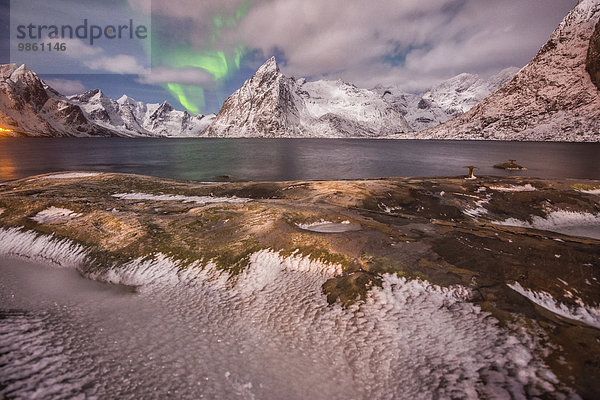 Aurora borealis mit Wolken  Felsen und Eis  kleine Stadt und Berge hinten  Insel Hamnoy  Lofoten  Norwegen  Europa