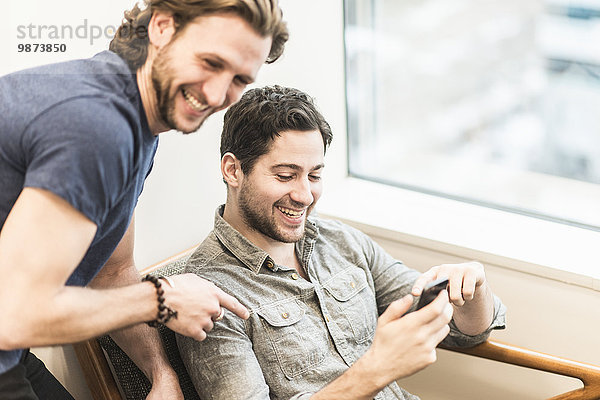 Ein Mann sitzt und schaut auf sein Smartphone und ein Kollege schaut ihm über die Schulter.