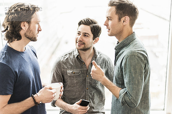 Drei Männer stehen und unterhalten sich  einer mit einer Tasse Kaffee  einer mit einem Smartphone.