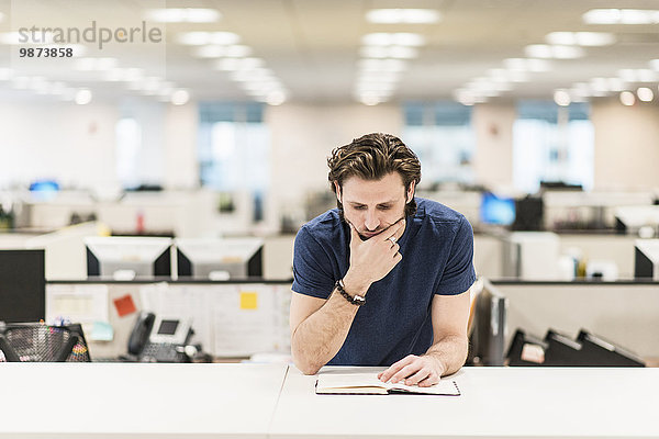 Ein Mann  der sich auf den Ellbogen lehnt und ein aufgeschlagenes Buch auf einem Büroschreibtisch betrachtet.