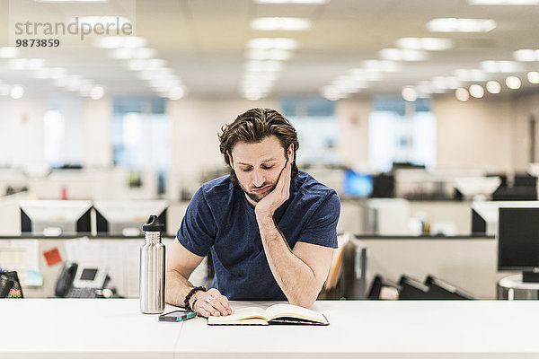 Ein Mann  der sich auf den Ellbogen lehnt und ein aufgeschlagenes Buch auf einem Büroschreibtisch betrachtet.