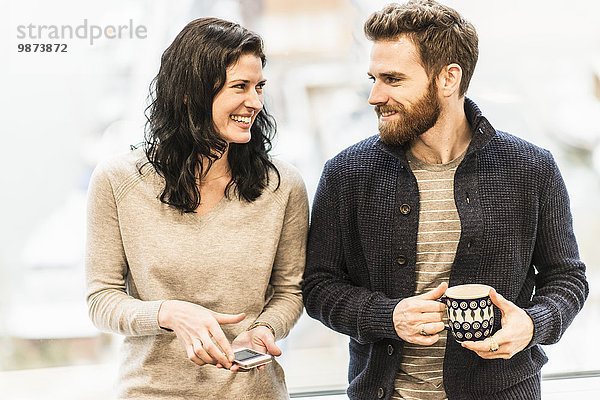 Eine Geschäftsfrau sitzt am Fenster  hält ein Smartphone in der Hand und unterhält sich mit einem Mann mit einer Kaffeetasse.