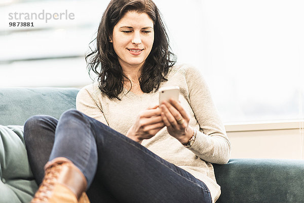 Eine Frau sitzt mit erhobenen Füßen auf einem Sofa und schaut auf ihr Smartphone.