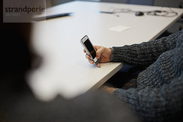 Ein Mann  der an einem Tisch in einem Büro sitzt und sein Smartphone benutzt.