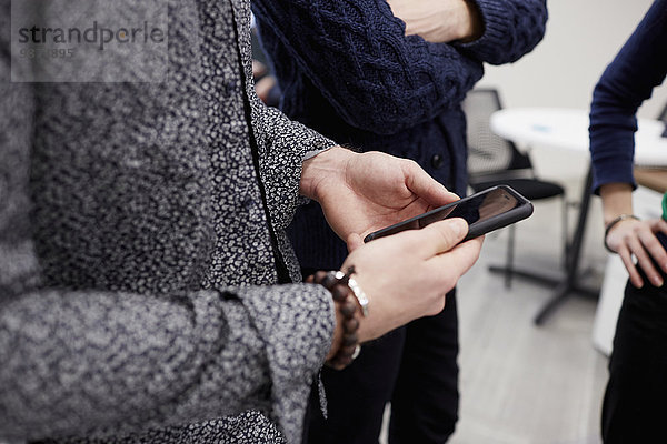 Drei Personen stehen in einem Büro  eine hält ein Smartphone in der Hand. Eine Besprechung im Stehen.