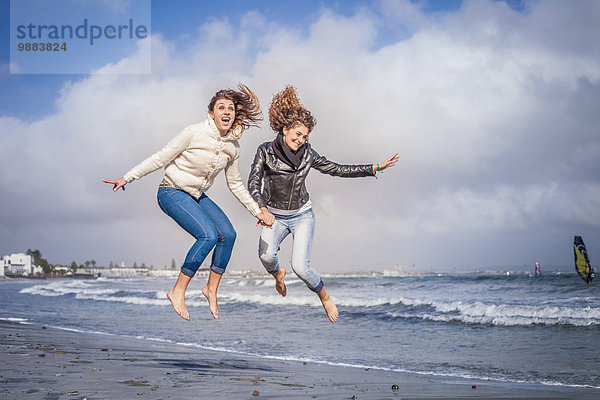 Zwei junge Frauen  die Händchen halten und am Strand springen.