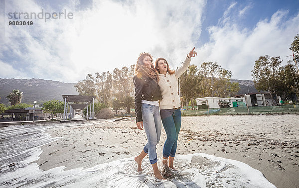 Zwei junge Frauen  die barfuß am Strand spazieren gehen.