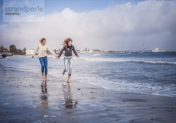 Zwei junge Frauen  die Hand in Hand am Strand laufen.