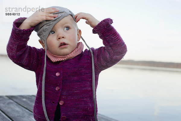 Kleines Mädchen auf dem Pier am See versucht  einen Hut aufzusetzen.