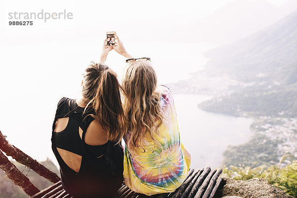 Rückansicht von zwei Freundinnen  die am Lake Atitlan  Guatemala  Smartphone Selfie nehmen