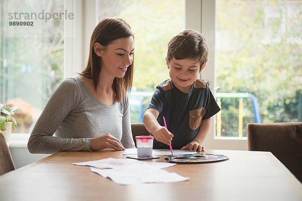 Junge lächelt mit Mutter und malt auf Papier am Tisch im Wohnzimmer.