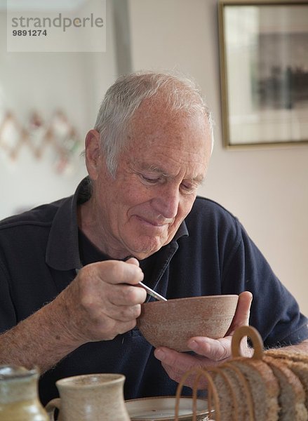 Älterer Mann  sitzt am Tisch  isst eine Schüssel mit Müsli.