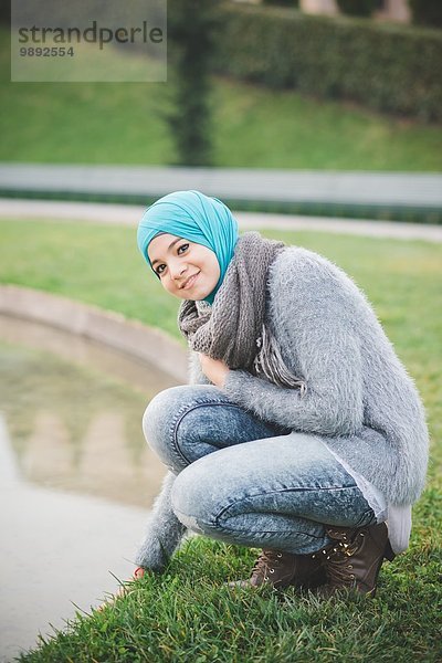 Porträt einer jungen Frau mit Hijab  die sich am Parksee hockt