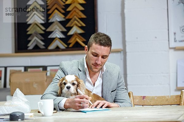 Mittlerer erwachsener Mann mit Hund beim Notieren am Schreibtisch in der Bilderrahmenwerkstatt