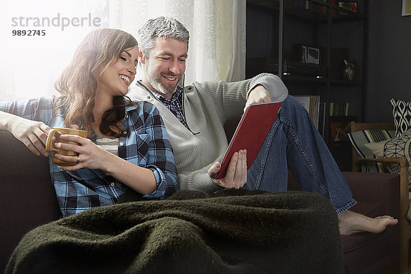 Junges Paar sitzt auf dem Sofa  trinkt Kaffee und schaut sich digitale Tabletten an.