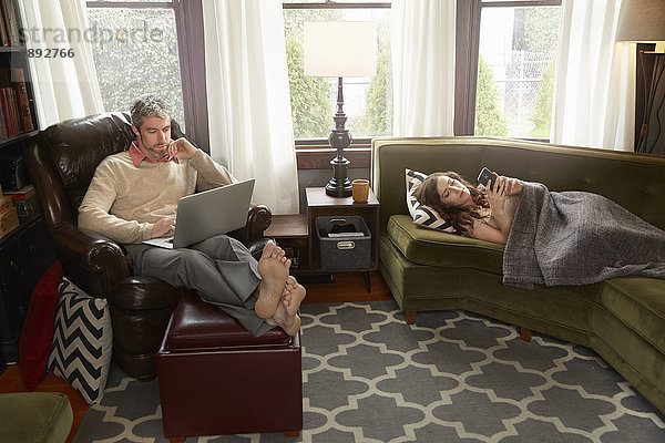 Junges Paar im Wohnzimmer mit Smartphone und Laptop