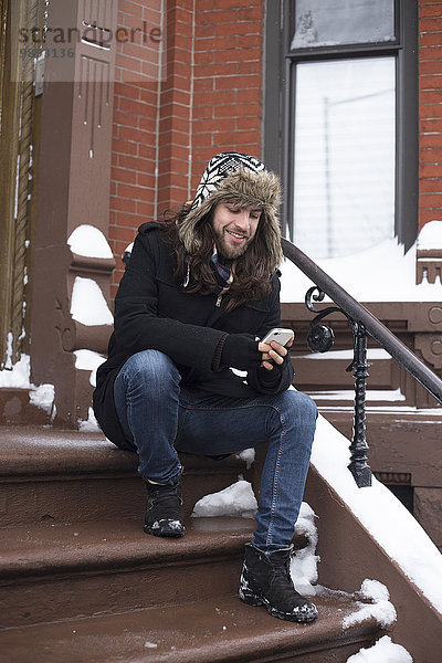 Junger Mann liest Text auf Smartphone auf schneebedeckter Haustürstufe